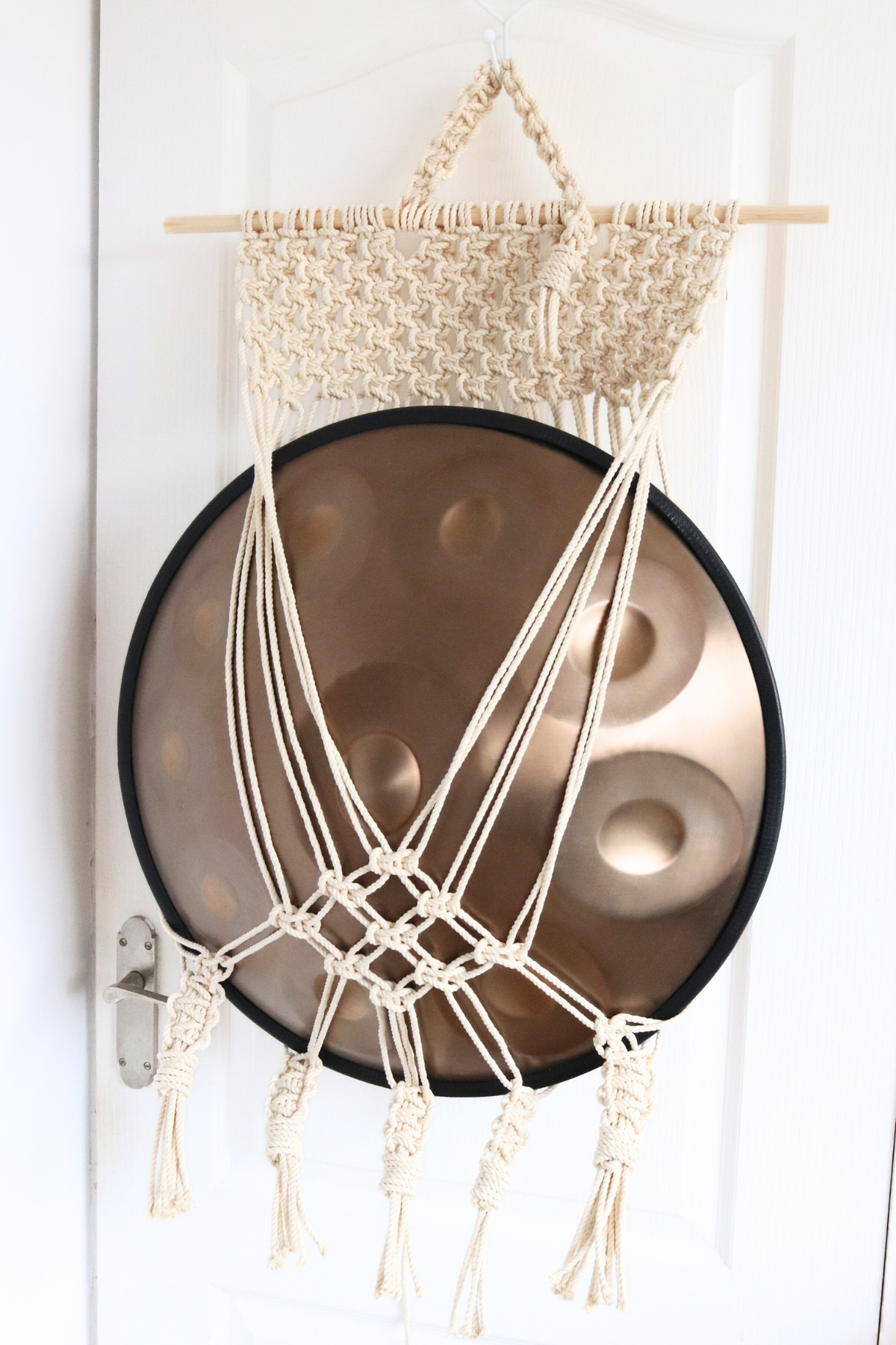Off-white macrame handpan hanger mount holding a bronze handpan on a pine dowel