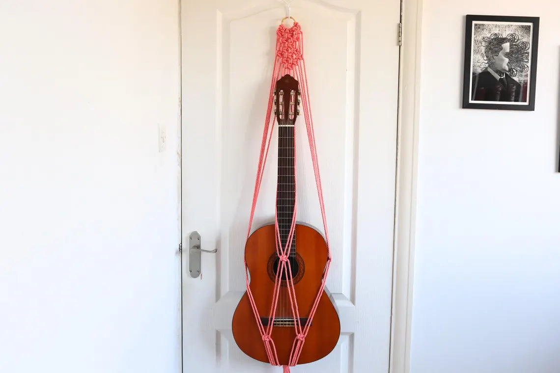 flamingo pink macrame guitar hanger with bamboo hoop holding an acoustic guitar on a wall