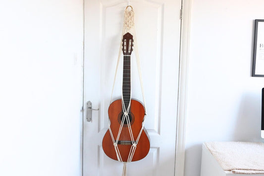 Off-white macrame guitar hanger with bamboo hoop holding an acoustic guitar on a wall
