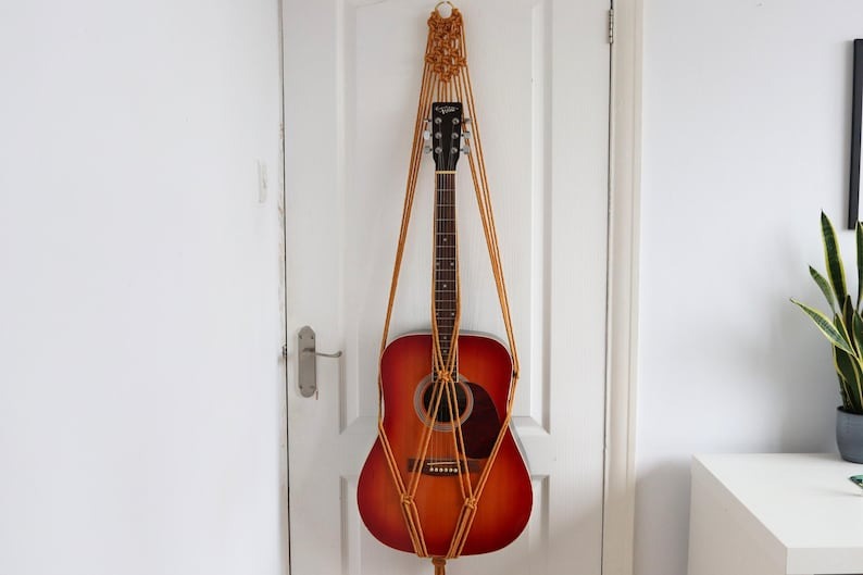mustard macrame guitar hanger with bamboo hoop holding an acoustic guitar on a wall