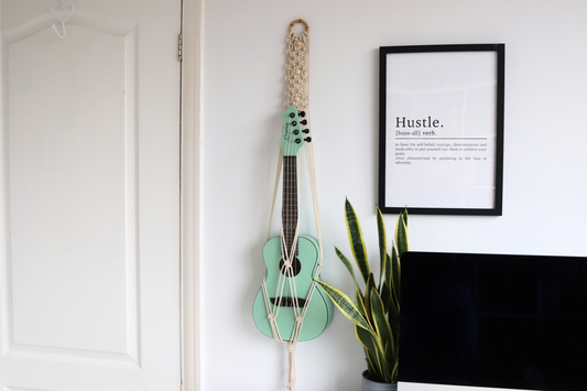 A green ukulele hanging on a wall mount with wooden beads, in front of a sign that says 'HUSTLE.' The mount is attached to a white door with a rope and a bamboo hoop.