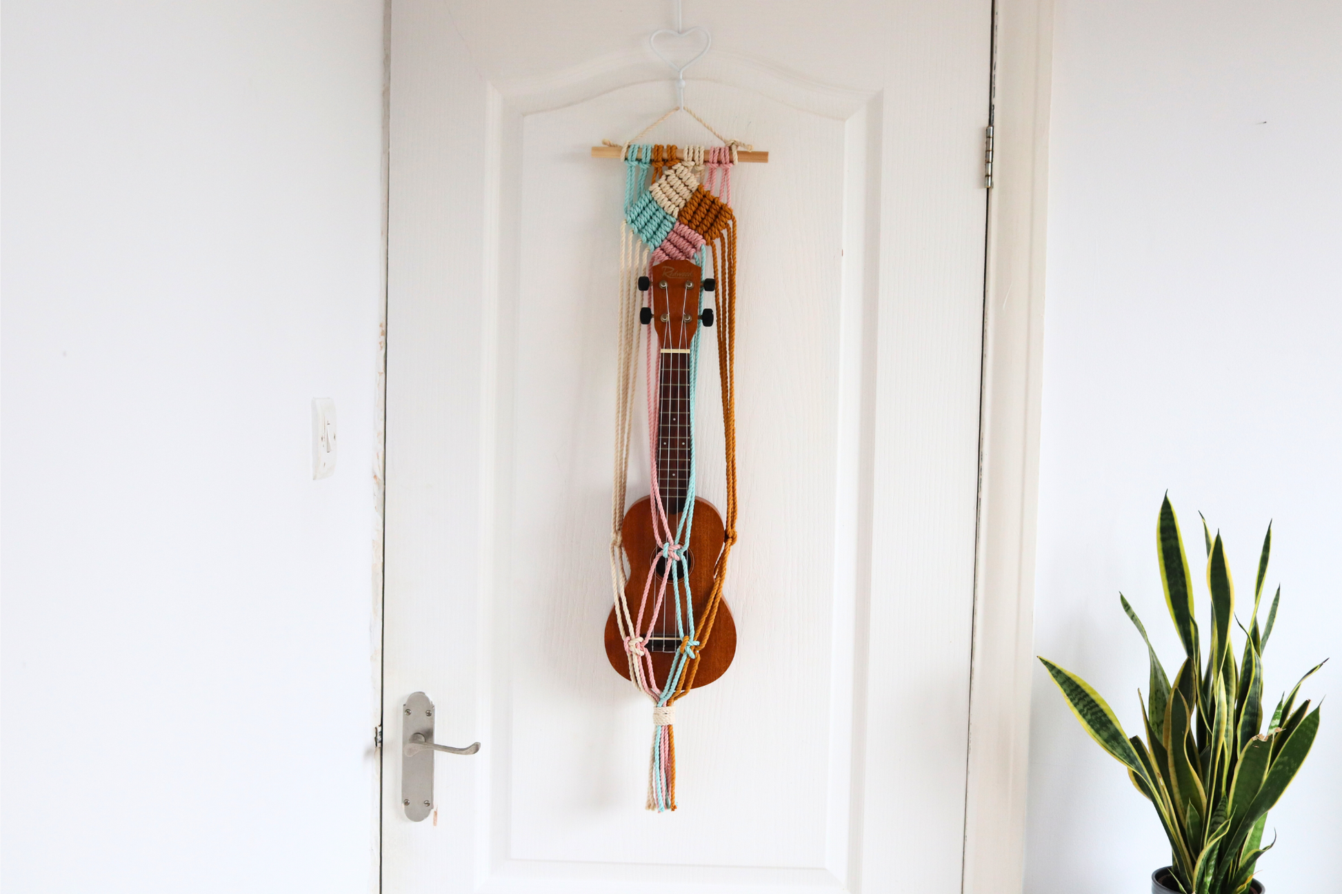 Handmade macrame ukulele wall mount in a multicolor 'Block Party' design, displaying a ukulele hanging on a white door.