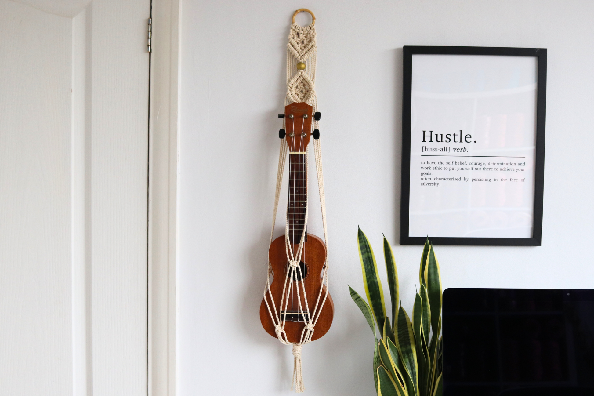 A brown ukulele hung on a white wall mount with gold beads, in a room with a black framed picture and a potted plant.